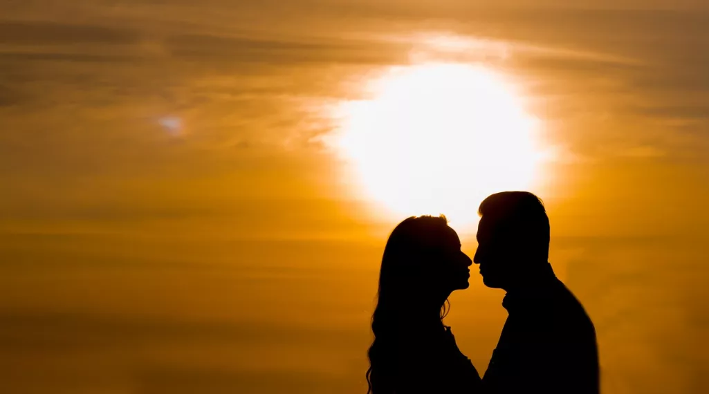 A couple leans in for a kiss in front of a setting sun.