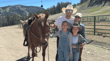 A smiling family of four, including John E., poses outdoors with a saddled brown horse at a mountain ranch, with pine-covered hills, fencing, and ski lift cables visible in the background on a sunny day.