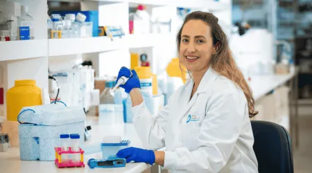 Dr. Mielke smiles while working in a research laboratory, wearing a white lab coat and blue gloves as she holds a pipette beside lab equipment.